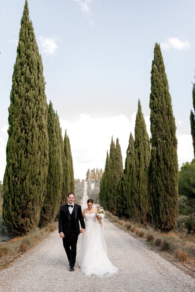 Hannah Chubb and Brandon Warner walking hand in hand along the cypress-lined driveway at Tenuta di Sticciano on their wedding day in Tuscany