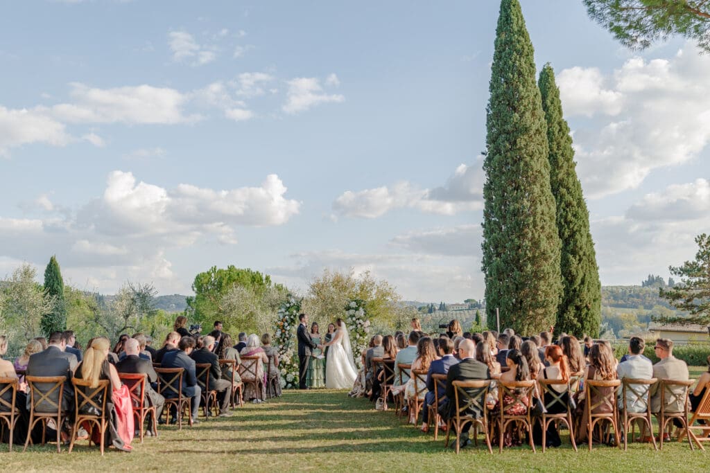 Wide view of Hannah and Brandon's outdoor wedding ceremony at Tenuta di Sticciano with cypress trees, guests seated on the lawn, and Tuscan hills beyond