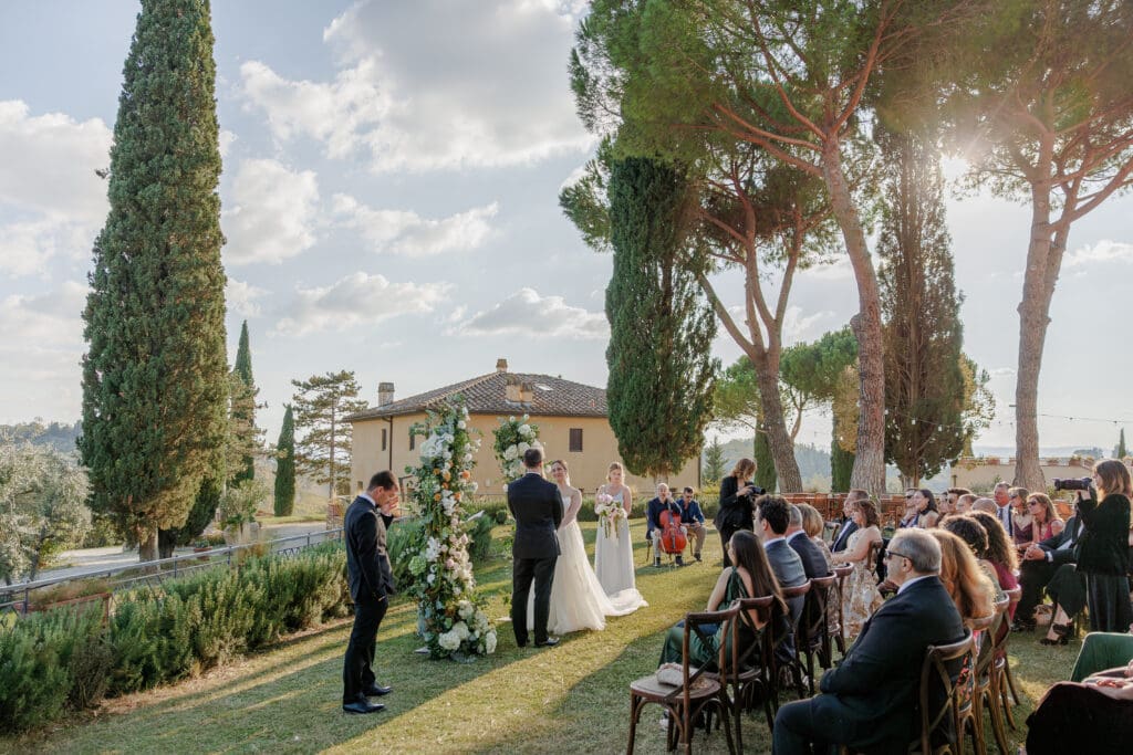 Hannah and Brandon exchanging vows at the floral arch at Tenuta di Sticciano with a cellist beside them and Tuscan estate buildings beyond