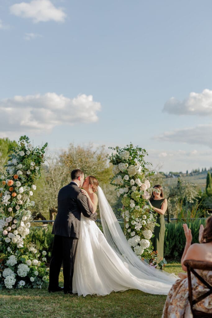Hannah and Brandon's first kiss beneath the white floral arch at their outdoor ceremony at Tenuta di Sticciano