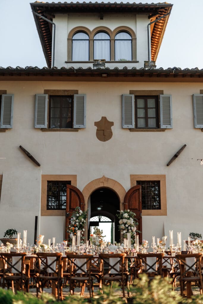 Long wedding reception table set in front of Tenuta di Sticciano villa facade with Violamalva floral columns flanking the stone arched doorway