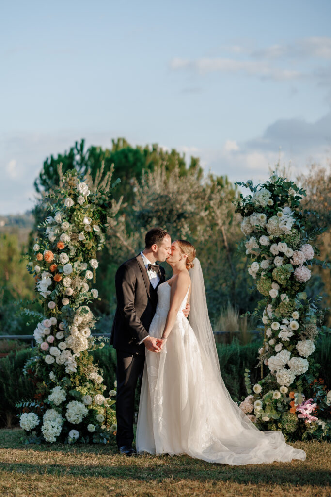 Hannah and Brandon kissing in front of the circular white floral arch with Tuscan countryside behind