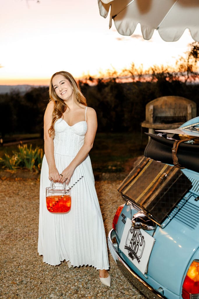 Bride Hannah in white dress holding an Aperol spritz beside a vintage Fiat 500 at sunset during a Tuscany wedding welcome party